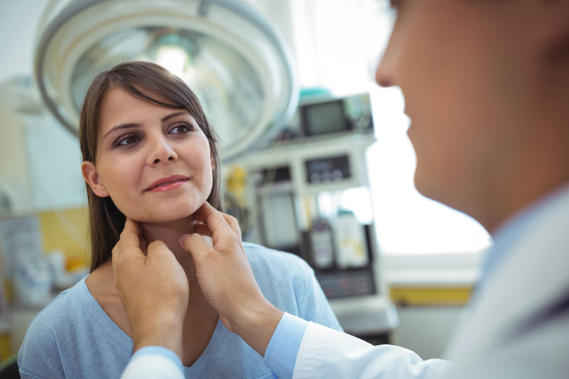 A doctor examines a smiling woman's thyroid and neck during a medical visit.