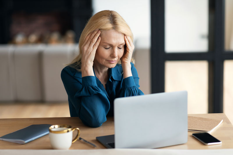 A woman in pain touches her head with both hands as she sits in front of a computer.