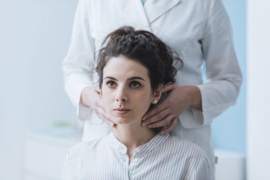 A doctor stands behind a seated woman and examines her thyroid and neck.